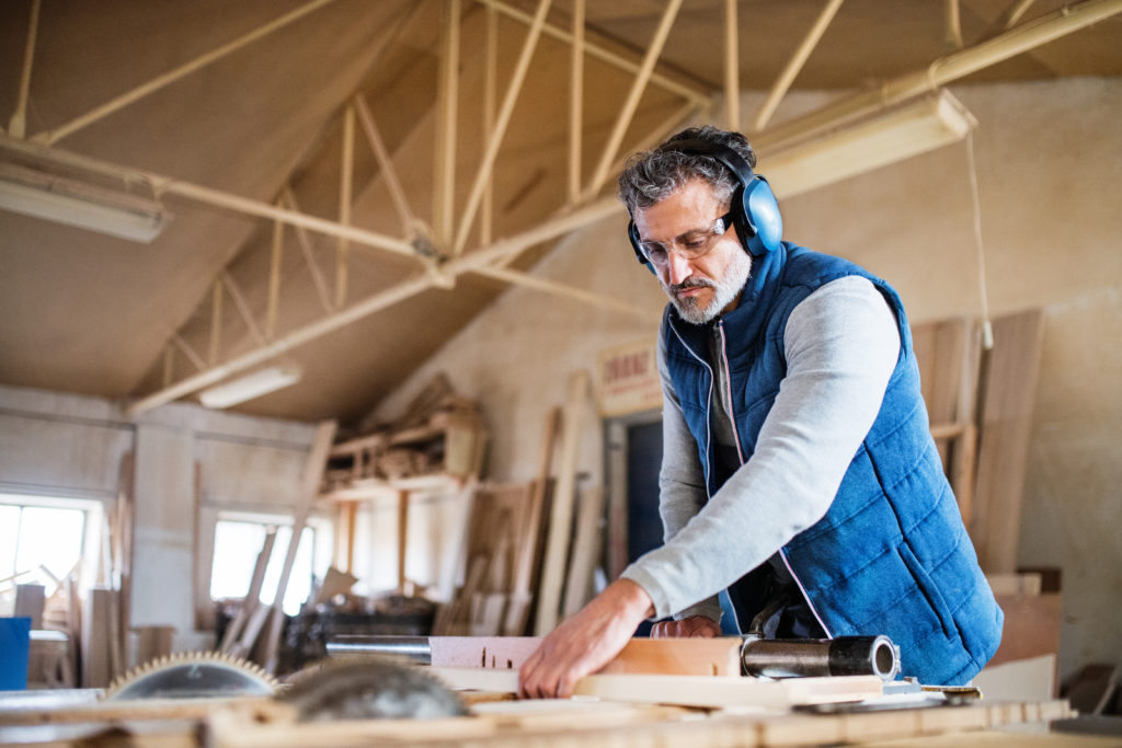 Mann in einer Holzwerkstatt mit Gehörschutzkopfhörern und Schutzbrille bei der Arbeit, Schutz vor berufsbedingtem Lärmschaden illustrierend.