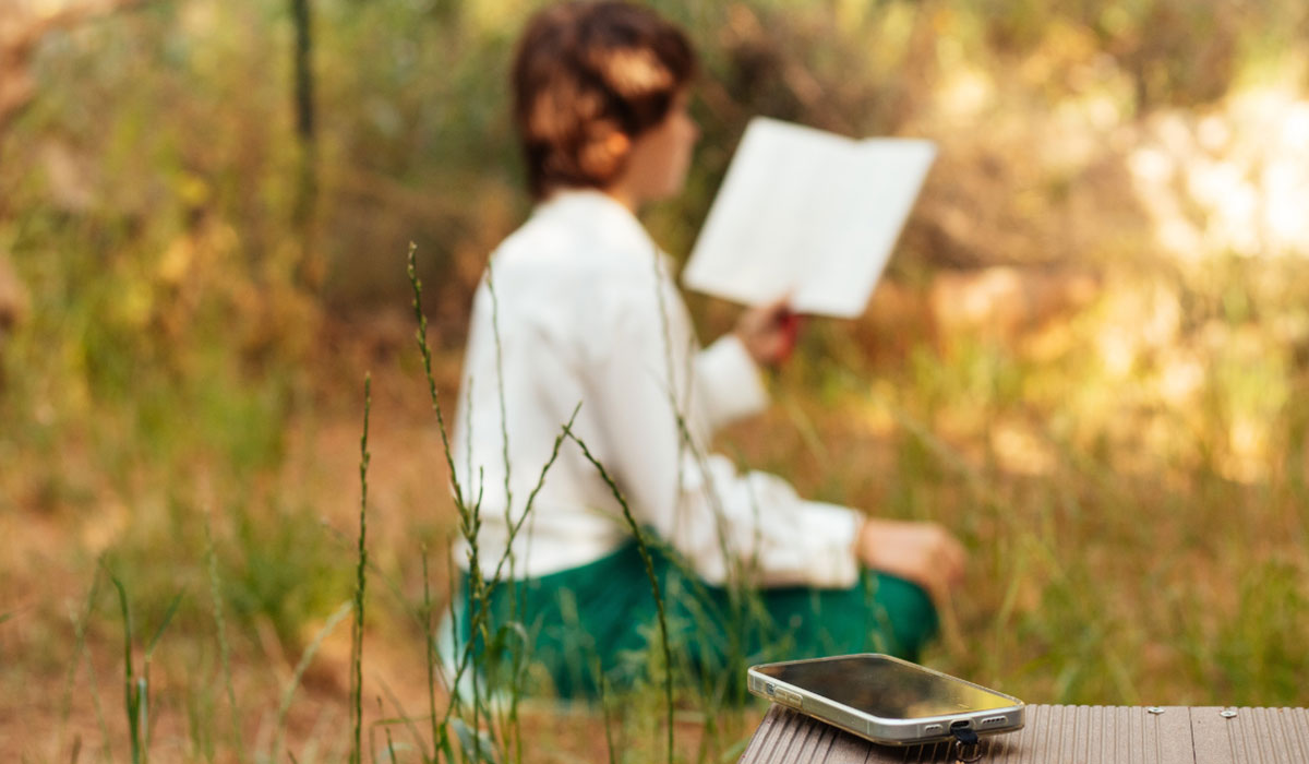 Smartphone-liegt-im-Vordergrund,-Person-liest-entspannt-im-Gruenen Smartphone-liegt-im-Vordergrund,-Person-liest-entspannt-im-Grünen